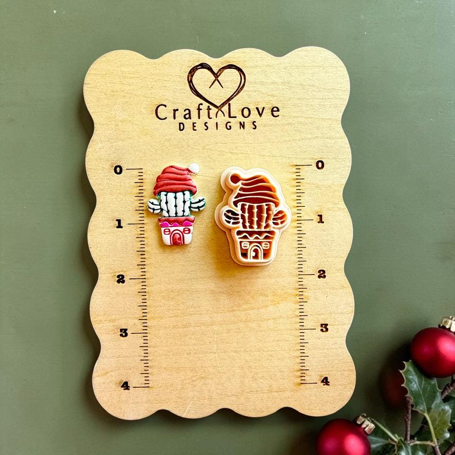 Finished Christmas cactus clay piece and matching polymer clay cutter displayed on a wooden Craft Love Designs sizing board, showing a potted cactus wearing a red Santa hat with Christmas ornaments and holly nearby.