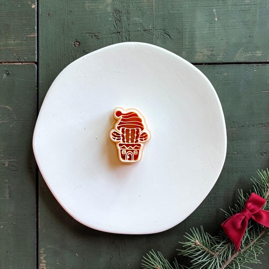 Christmas cactus polymer clay cutter centered on a white plate, showing a potted cactus with raised arms, decorative pot, and floppy Santa hat against a dark green wood background.