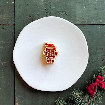 Christmas cactus polymer clay cutter centered on a white plate, showing a potted cactus with raised arms, decorative pot, and floppy Santa hat against a dark green wood background.