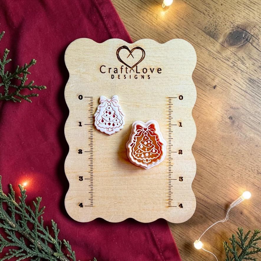 Finished white Christmas tree clay piece with red decorative accents and a bow beside the matching polymer clay cutter on a wooden Craft Love Designs sizing board with red fabric, evergreen branches, and warm holiday lights.