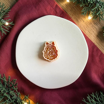 Bow Christmas tree polymer clay cutter centered on a white plate, showing layered scalloped tree branches, decorative details, and a large bow at the top against a festive red and evergreen background.