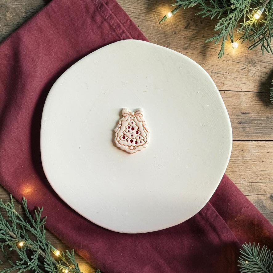 Finished white Christmas tree clay piece centered on a white plate, featuring a large bow at the top, layered scalloped branches, and red ornament-style details with holiday greenery and lights around the scene.