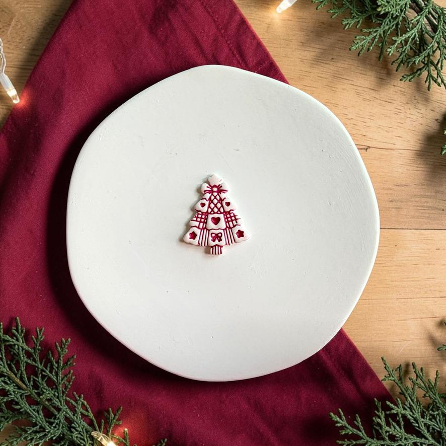 Finished white Christmas tree clay piece centered on a white plate, featuring dark red hearts, bows, stripes, lattice patterns, and decorative holiday details against a red fabric and evergreen background.