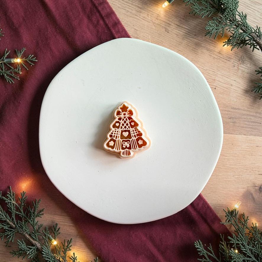 Decorative Christmas tree polymer clay cutter centered on a white plate, showing the layered tree silhouette with hearts, bow, lattice patterns, stripes, and small ornamental details.