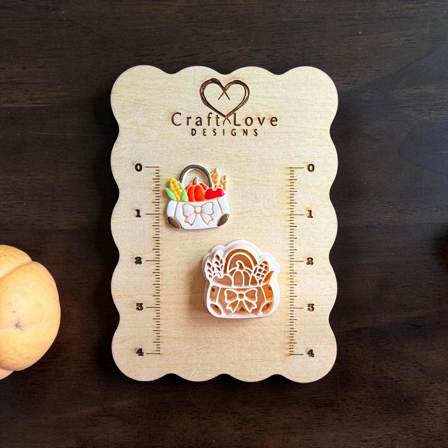 Finished fall harvest market bag clay piece beside the matching polymer clay cutter on a wooden Craft Love Designs sizing board, showing a tote filled with pumpkin, corn, wheat, and vegetables with a bow on the front.