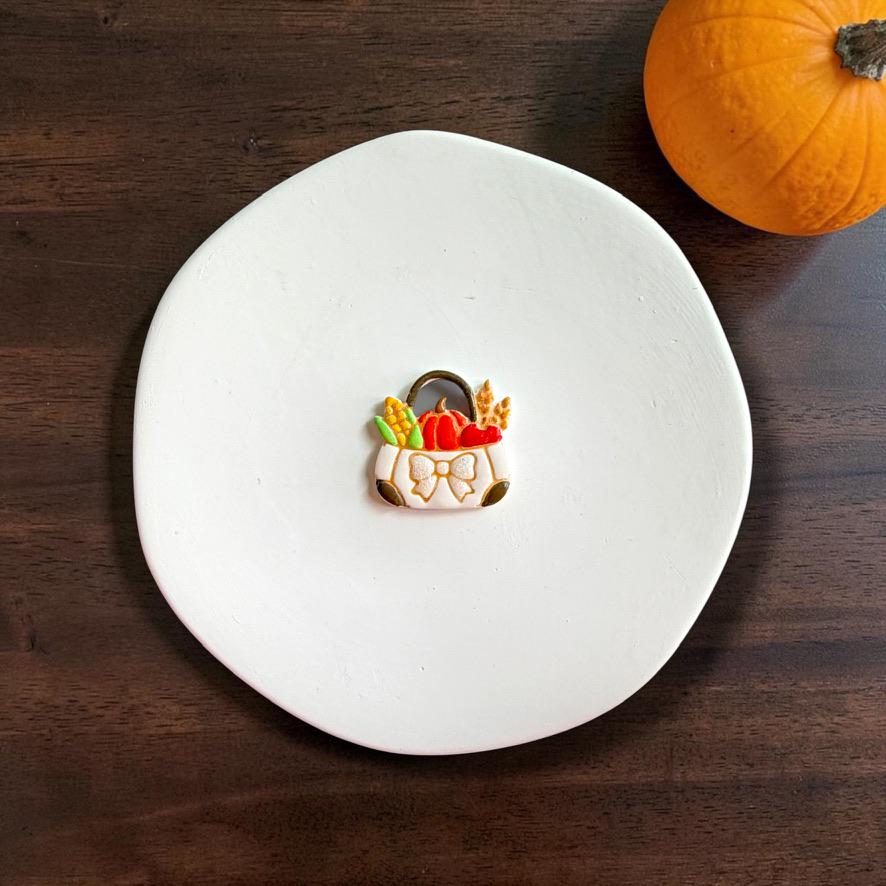 Finished fall harvest market bag clay piece centered on a white plate, featuring a brown handle, orange pumpkin, yellow corn, wheat, green accents, and a white bow on the front.