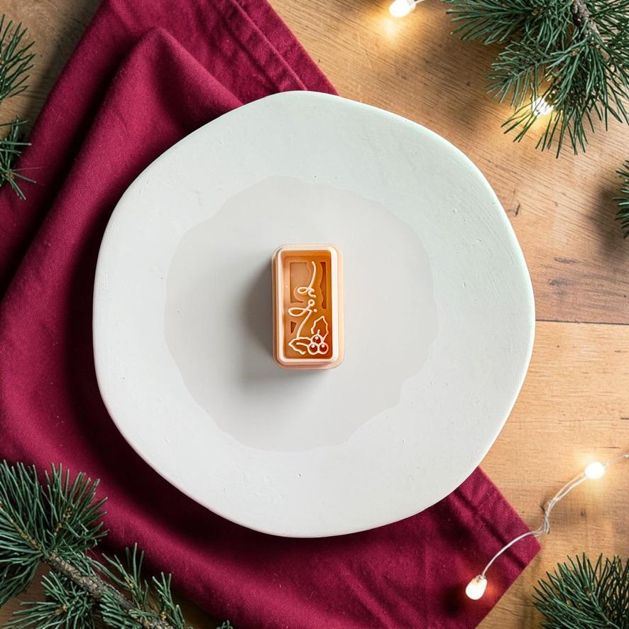 Joy and holly polymer clay cutter centered on a white plate, showing the slim rectangular shape with script lettering and holly leaves with berry details.
