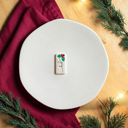 Finished white Joy clay piece centered on a white plate, featuring black script lettering with green holly leaves and red berries at the top against a festive red and evergreen background.