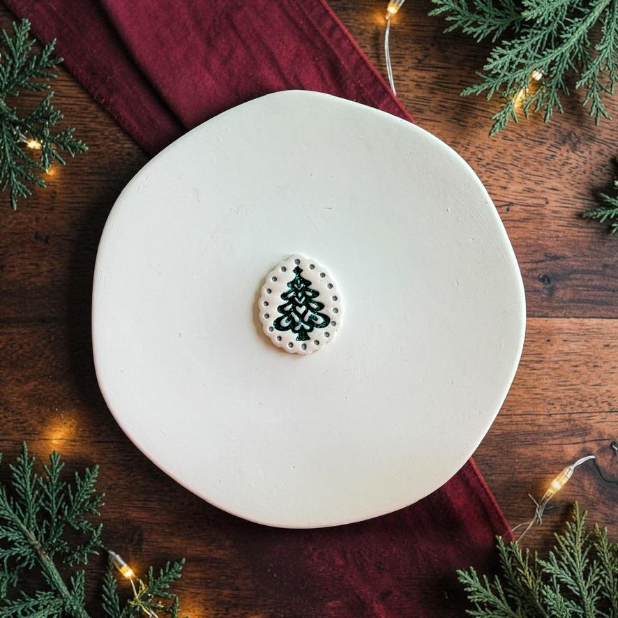 Finished white scalloped oval clay piece centered on a white plate, featuring a dark green stylized Christmas tree surrounded by a dotted border with festive greenery and lights in the background.
Photo 3: