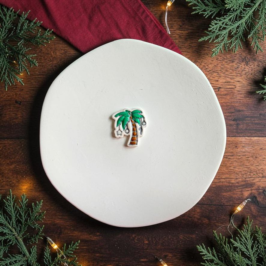 Finished Christmas palm tree clay piece centered on a white plate, featuring green palm leaves, a brown decorated trunk, and silver hanging ornaments against a dark wood holiday background.