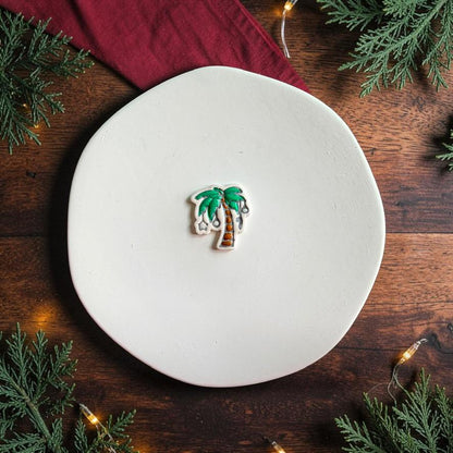 Finished Christmas palm tree clay piece centered on a white plate, featuring green palm leaves, a brown decorated trunk, and silver hanging ornaments against a dark wood holiday background.