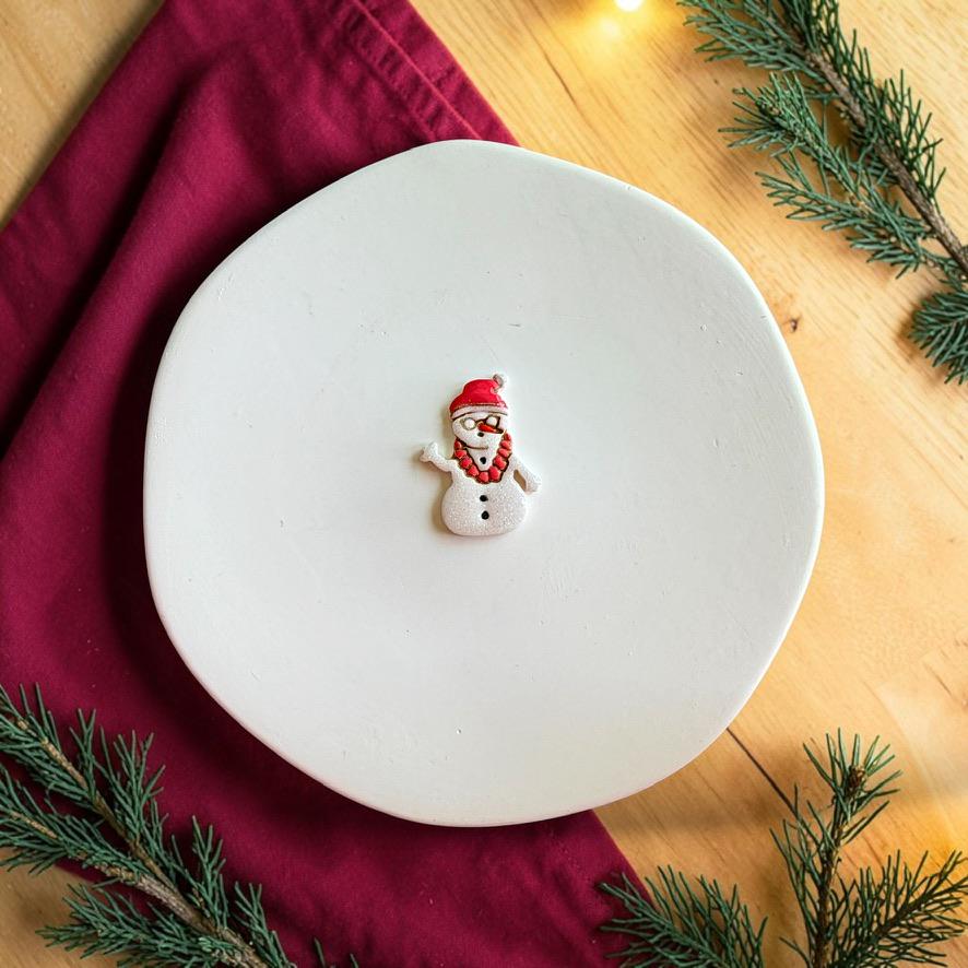 Finished tropical snowman clay piece centered on a white plate, showing a white snowman with a red Santa hat, sunglasses, lei, and black buttons.