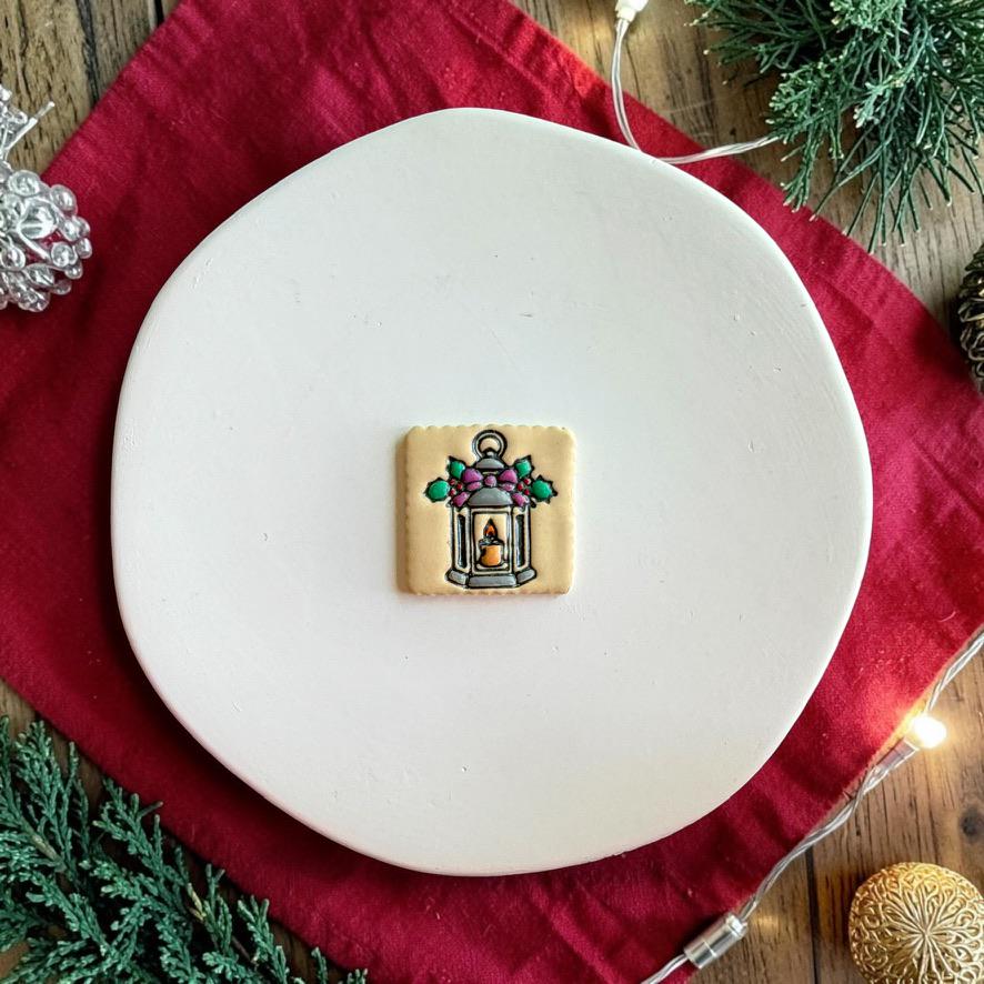 Finished square Christmas lantern clay piece centered on a white plate, featuring a silver and blue lantern with a glowing candle, purple bow, green holly leaves, and colorful berry accents.