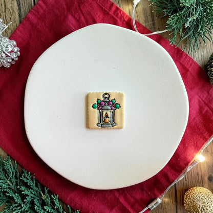 Finished square Christmas lantern clay piece centered on a white plate, featuring a silver and blue lantern with a glowing candle, purple bow, green holly leaves, and colorful berry accents.