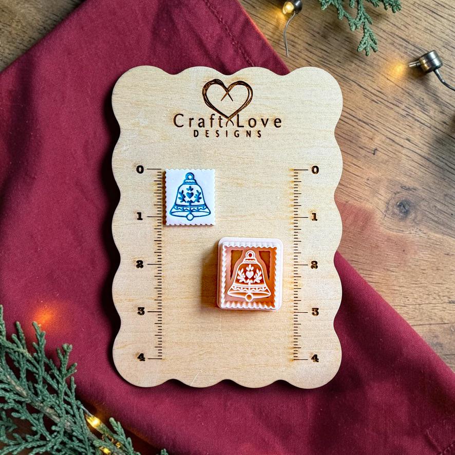Finished white square clay piece with a blue decorative Christmas bell beside the matching polymer clay cutter on a wooden Craft Love Designs sizing board with red fabric, evergreen branches, and holiday lights.