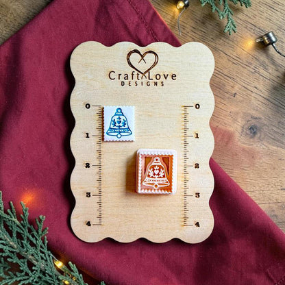 Finished white square clay piece with a blue decorative Christmas bell beside the matching polymer clay cutter on a wooden Craft Love Designs sizing board with red fabric, evergreen branches, and holiday lights.