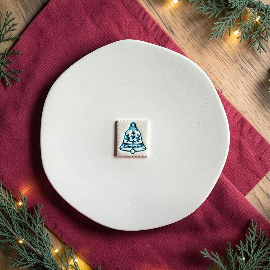 Finished white square clay piece centered on a white plate, featuring a blue folk-art Christmas bell with heart and leafy decorative details.