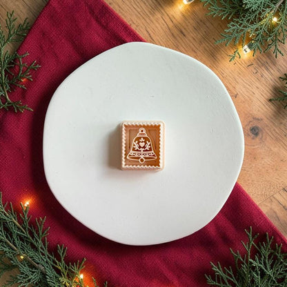 Christmas bell polymer clay cutter centered on a white plate, showing the bell shape with heart, floral, leafy, and ornamental details inside a scalloped square border.