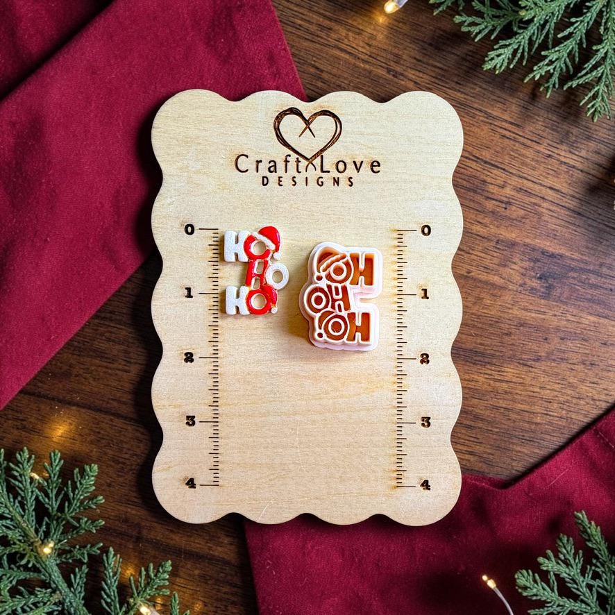 Finished white and red Ho Ho Ho clay piece beside the matching polymer clay cutter on a wooden Craft Love Designs sizing board with red fabric, greenery, and holiday lights.